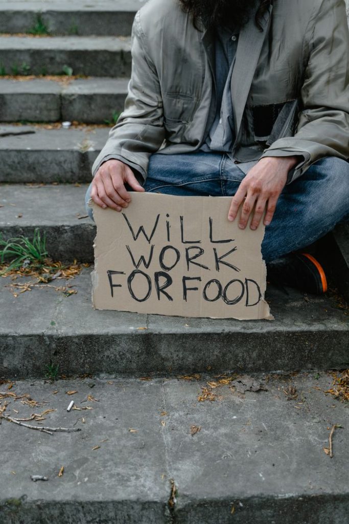 An individual sitting on stairs, holding a sign reading 'Will Work for Food', depicting economic struggle.