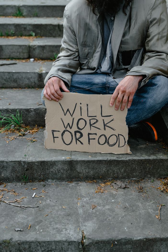 An individual sitting on stairs, holding a sign reading 'Will Work for Food', depicting economic struggle.